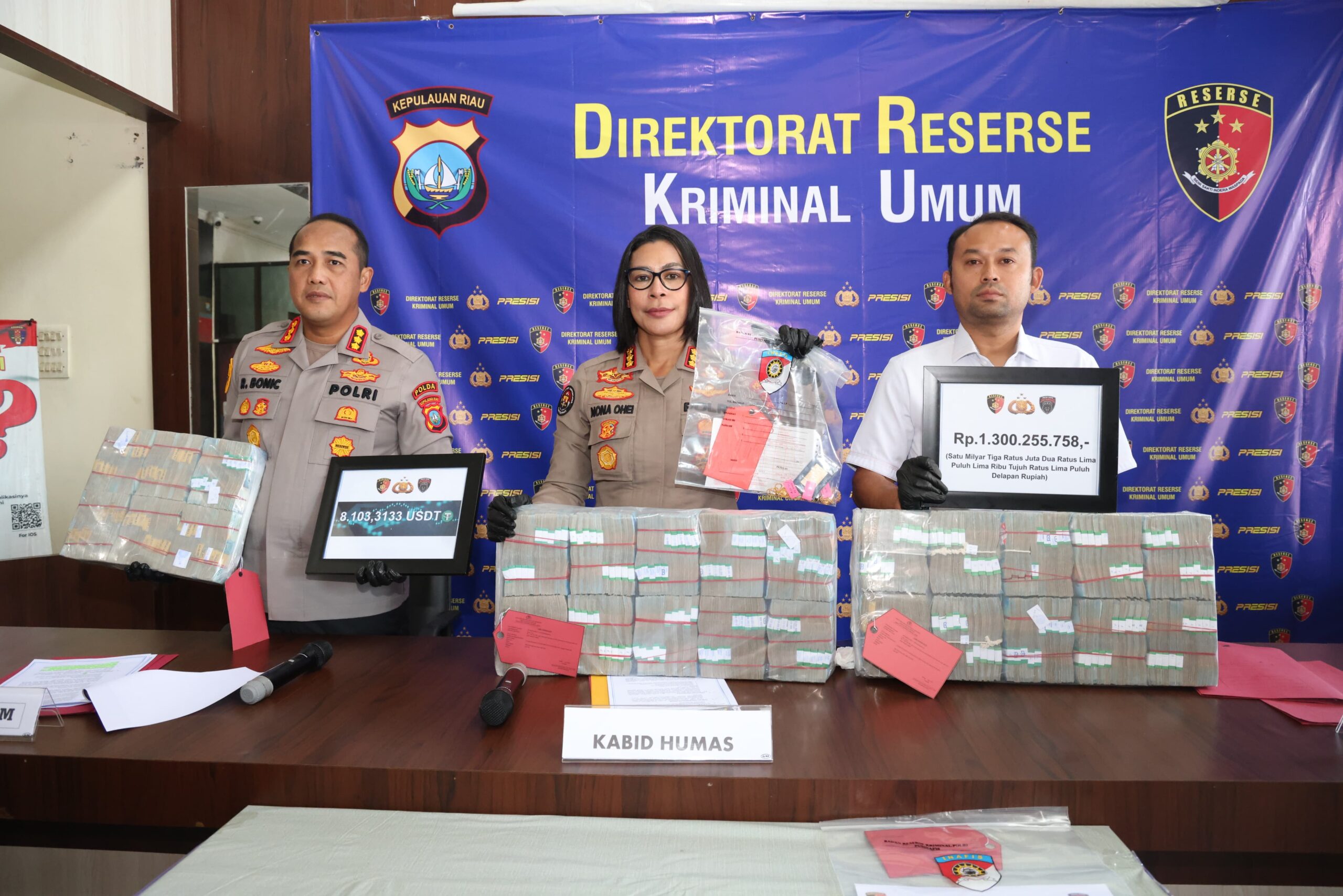 Tiga pejabat Polda Kepri berdiri di depan tumpukan uang sitaan, barang bukti kripto, dan papan nominal rupiah saat pengungkapan kasus judi online.